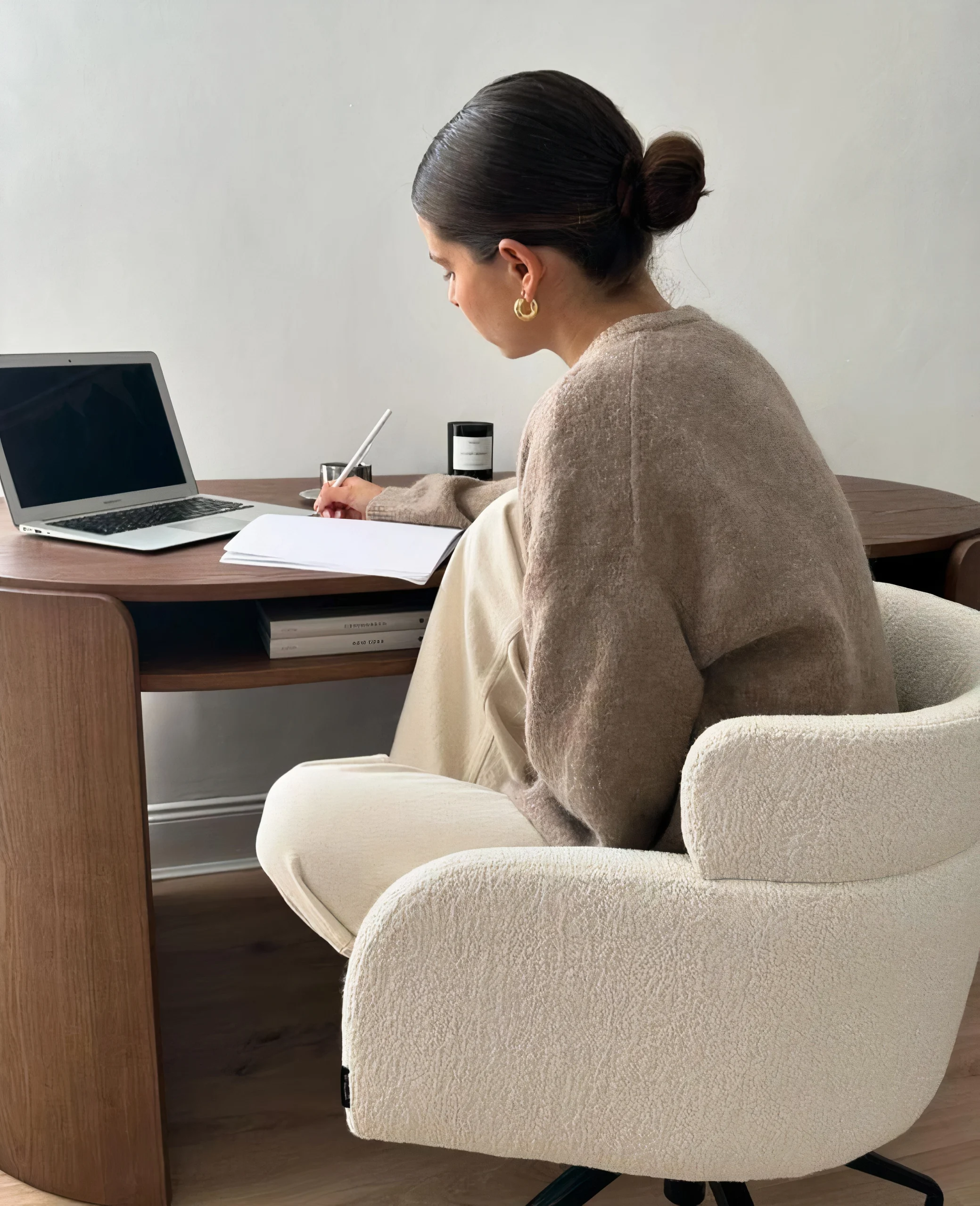 Jeune fille de 3/4 avec petit chignon, écrivant sur son cahier devant son ordinateur dans un décor beige et bois